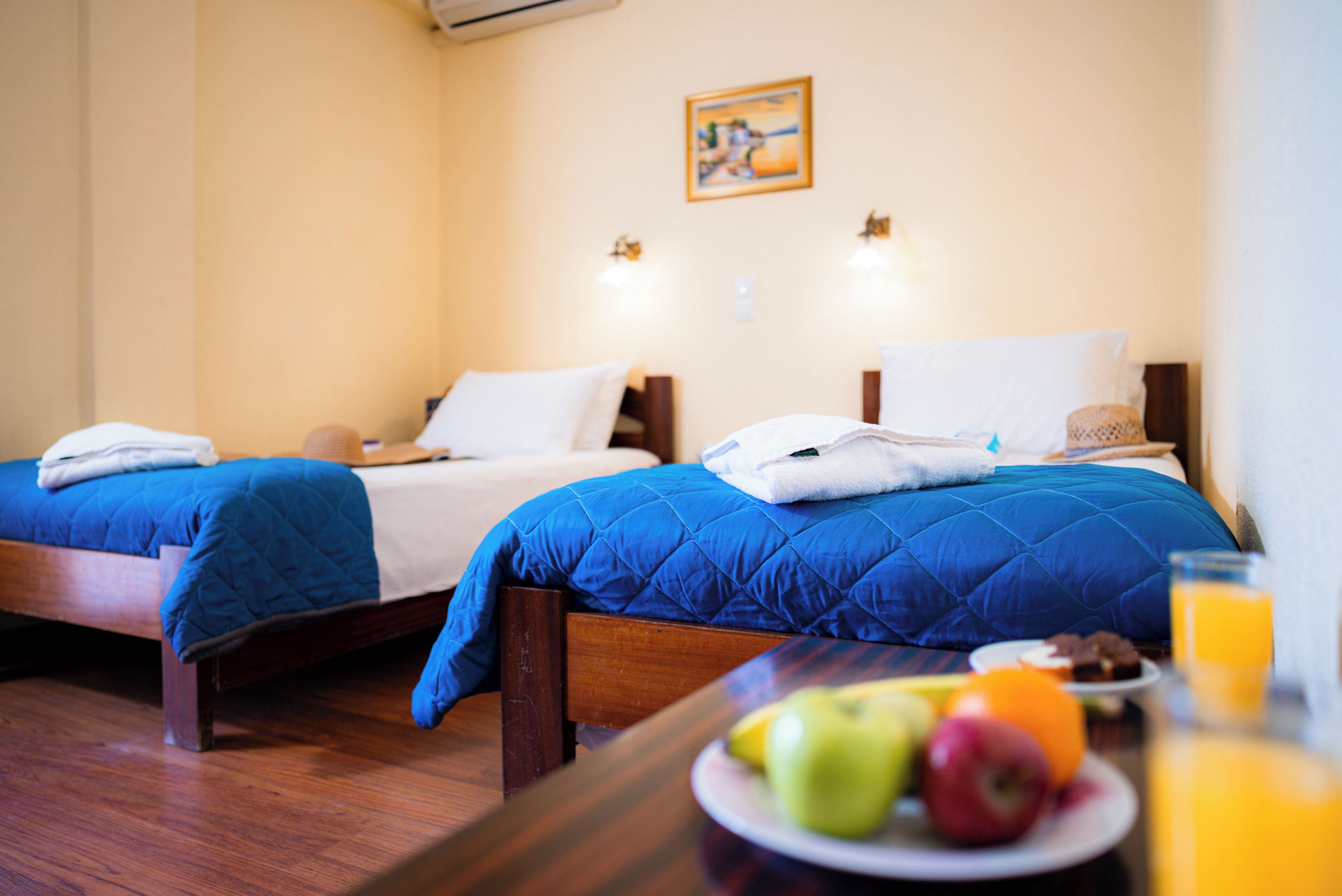 A hotel room with two beds, a wooden floor, a dining table with fruit and a glass of orange juice, and a painting on the wall.