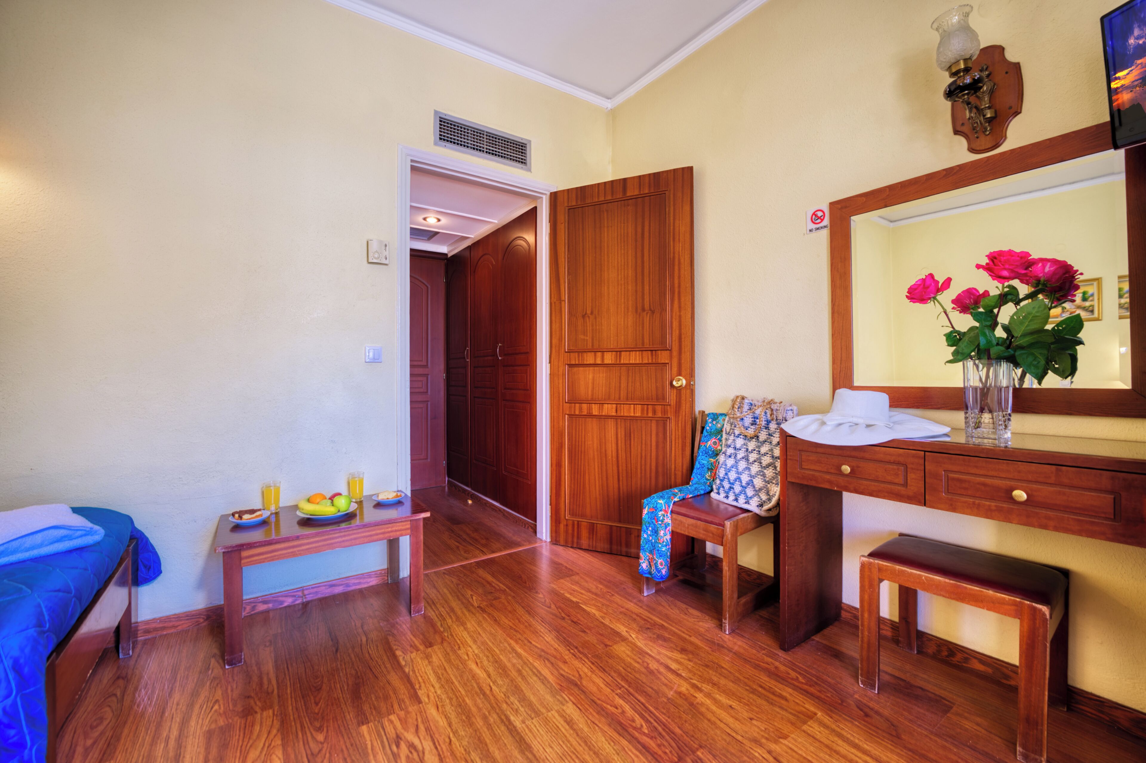 A room with a bed, a wooden desk, a chair, a small table with fruit, a mirror, and a door leading to another room.