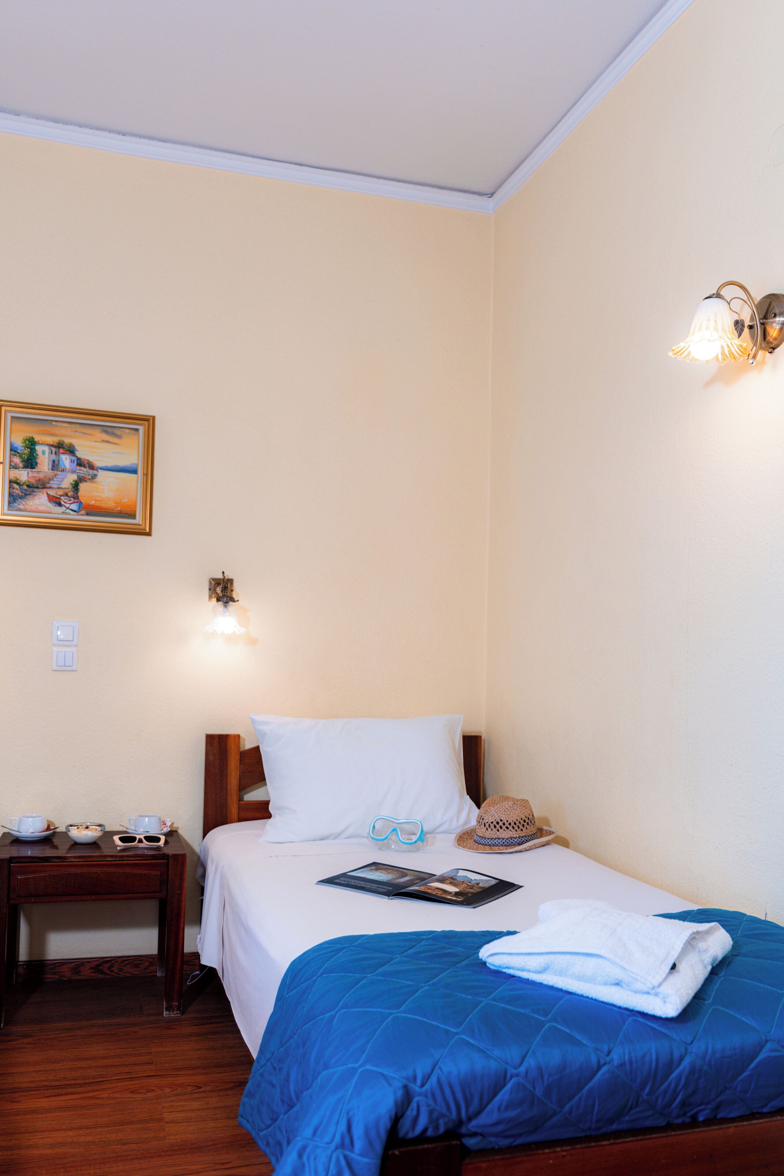 Una habitación de hotel con una cama, una mesita de noche, una lámpara, un cuadro y un sombrero sobre la cama.