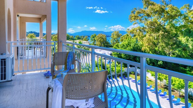 A balcony with a view of a pool and mountains, a chair, and a table with a towel. - Sunset Hotel Corfu (Corfu)
