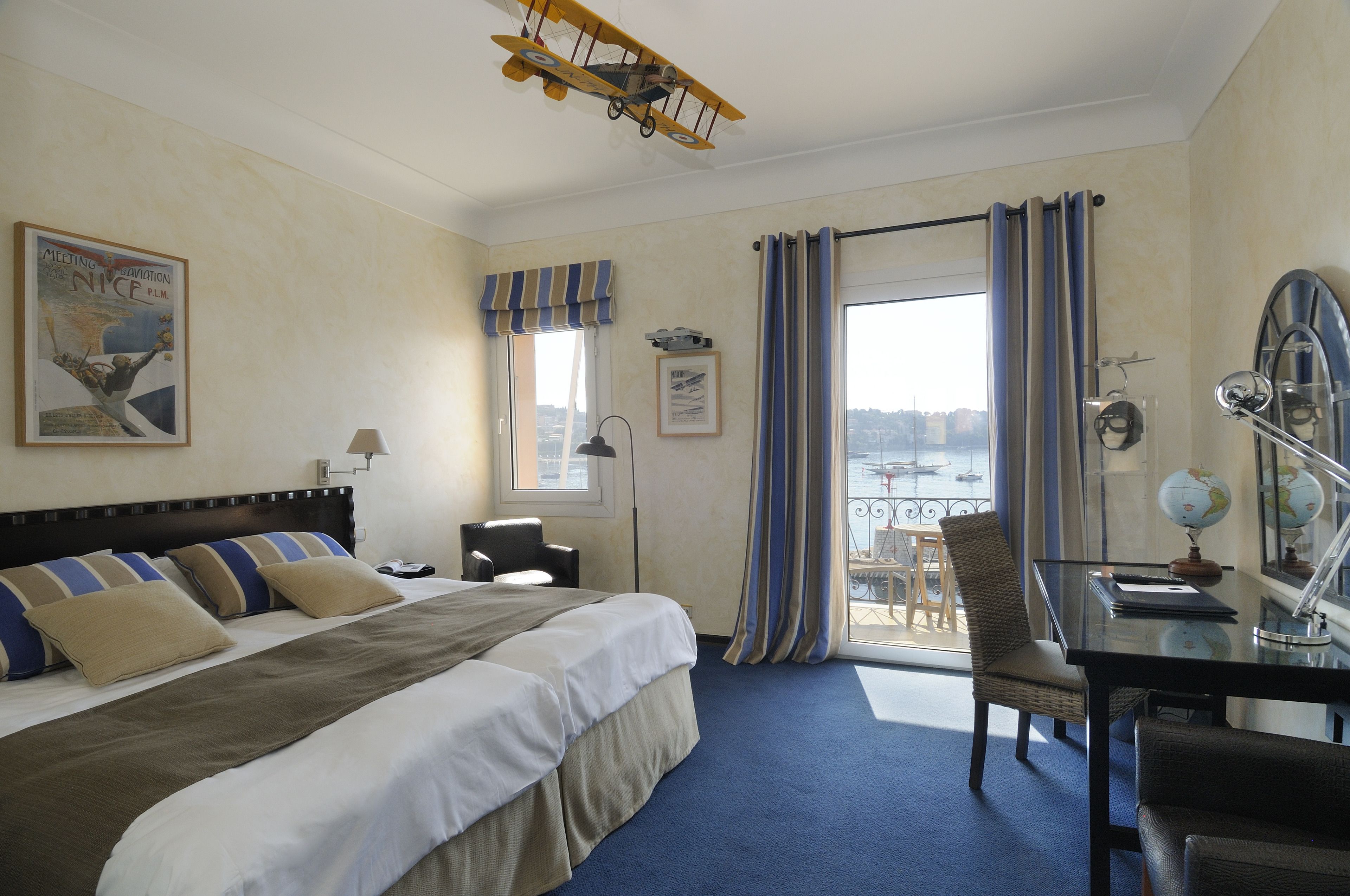 A hotel room with a bed, a desk with a globe, a chair, a window with curtains, and a ceiling airplane model.