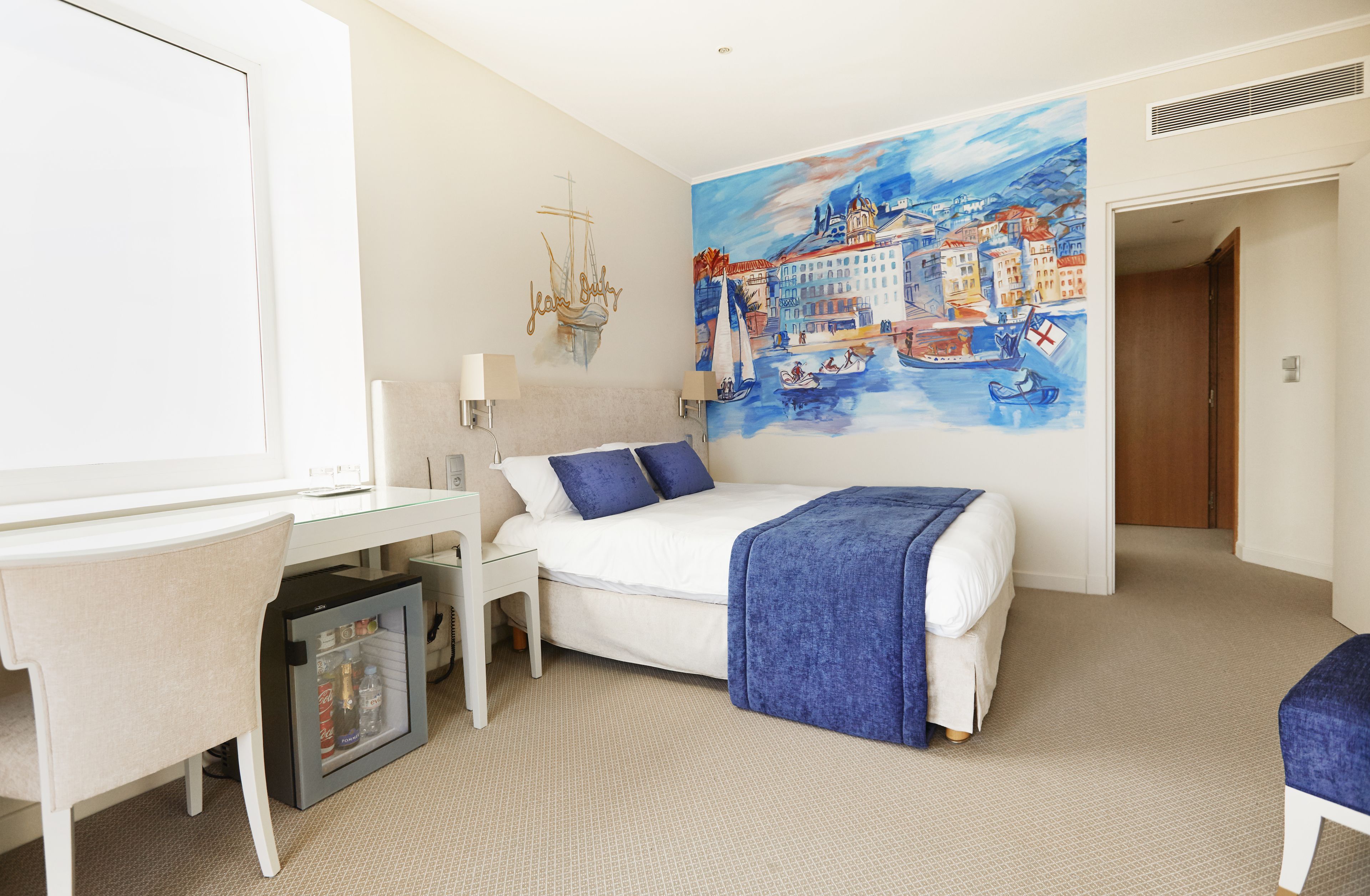 A hotel room with a bed, a desk, a chair, a small refrigerator, and a mural of a coastal town.