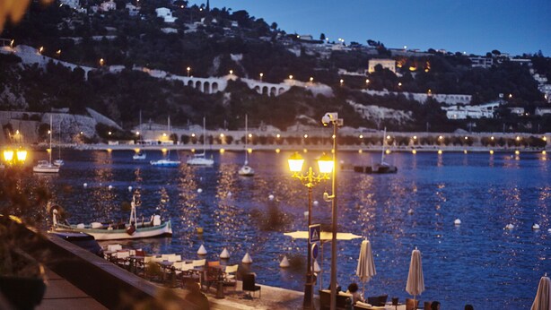 A waterfront restaurant with outdoor seating overlooking a body of water, boats, and a hillside in the background. - Welcome Hôtel (Villefranche-sur-Mer)