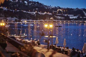 A waterfront restaurant with outdoor seating overlooking a body of water, boats, and a hillside in the background.