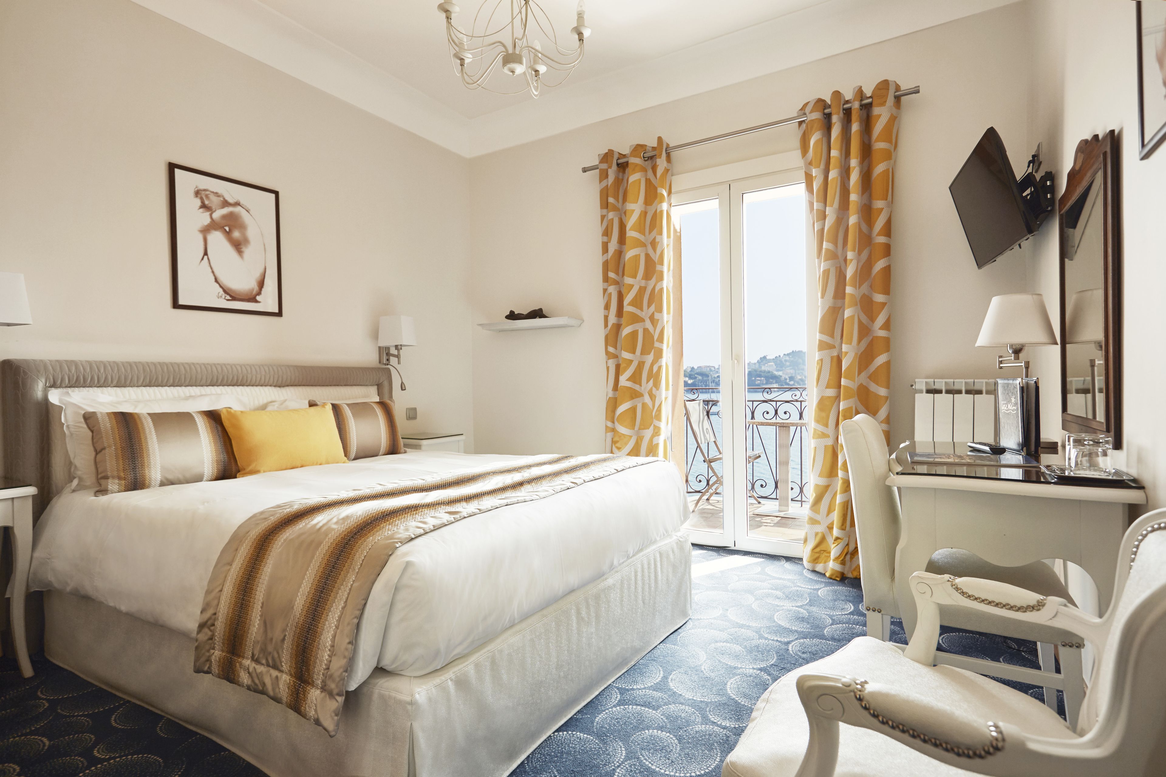 A hotel room with a large bed, a desk with a chair, a balcony with curtains, and a city view.