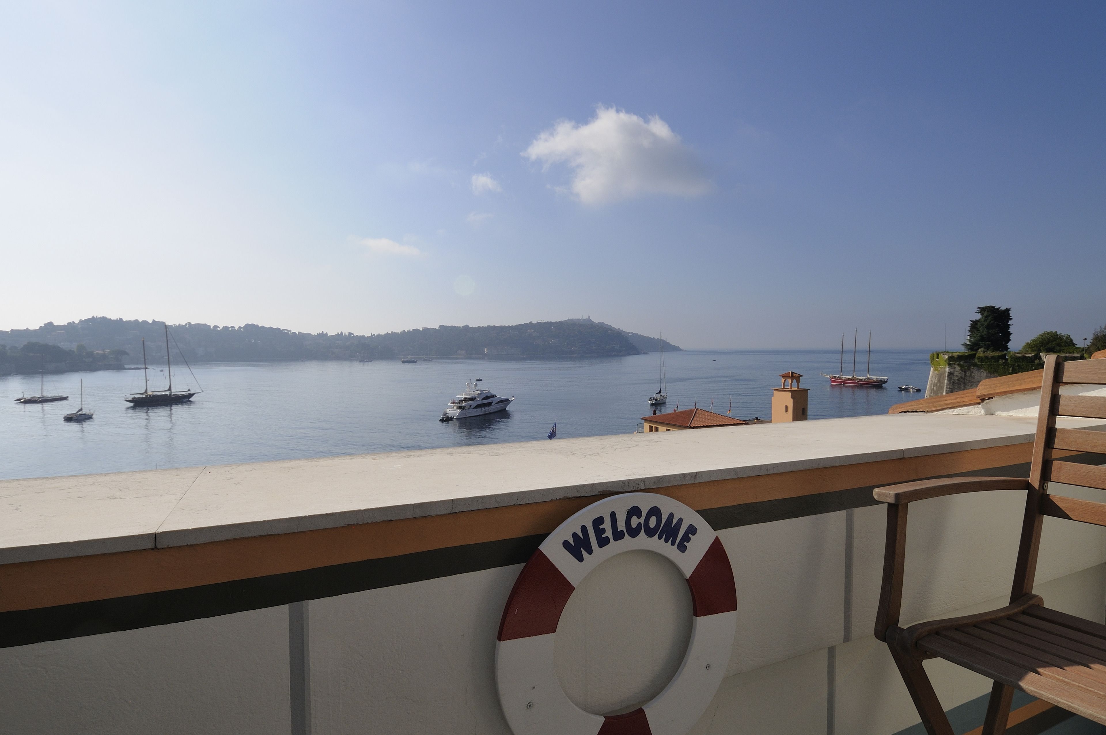 A balcony with a "WELCOME" sign, overlooking calm waters with boats and a distant hill.