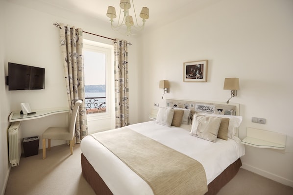A hotel room with a large bed, a desk, a chair, a TV, and a view of the sea.