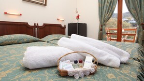A hotel room with a bed, a wooden headboard, a painting on the wall, a window with curtains, a view of the outside, and a basket with toiletries.