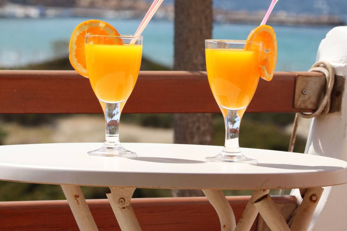 Two glasses of orange juice on a white table with a view of the sea and a tree in the background.