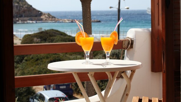 Two glasses of orange juice on a small white table with a view of the sea and a tree in the background.