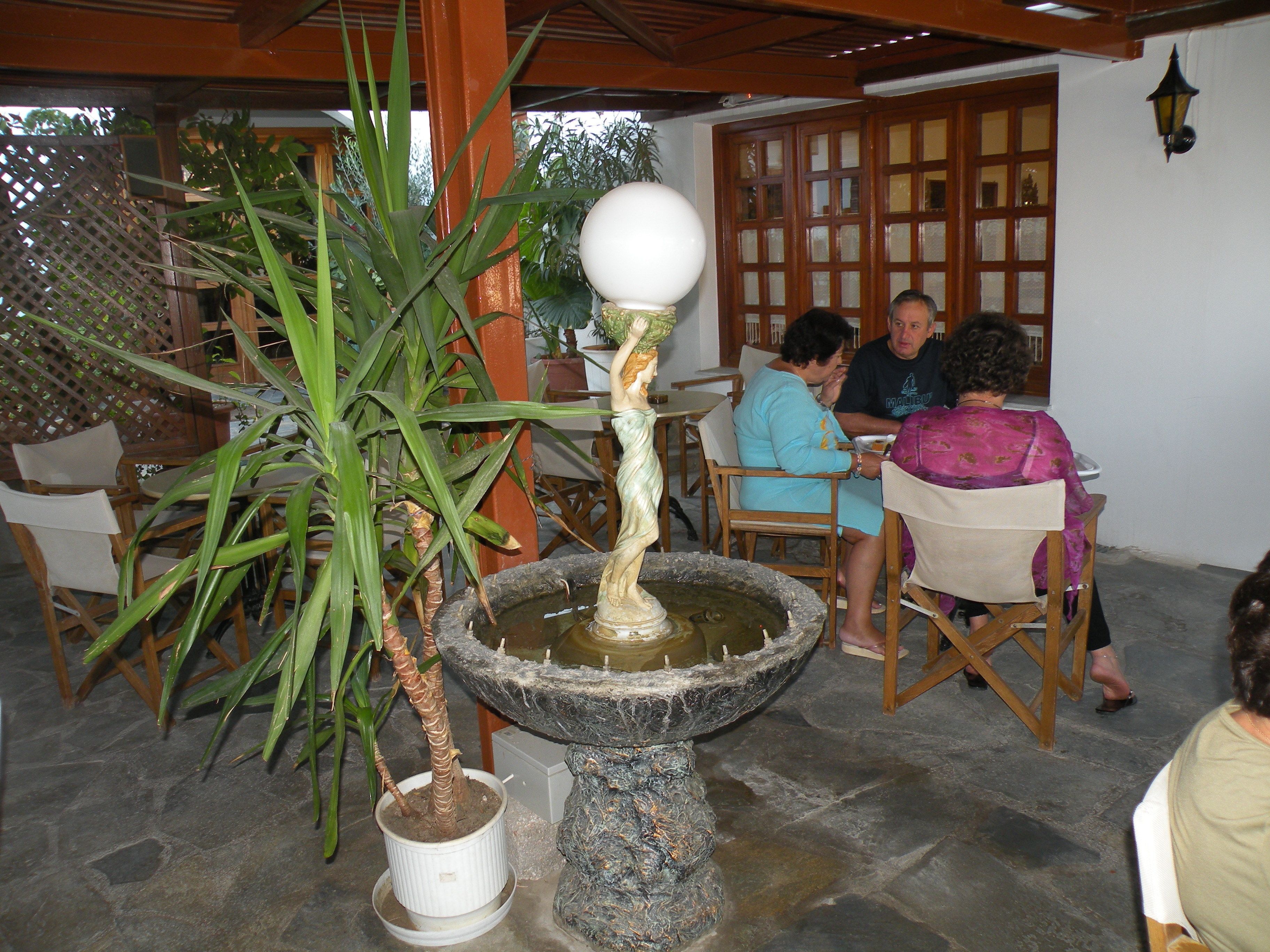 A stone fountain with a spherical top, surrounded by potted plants and wooden chairs.
