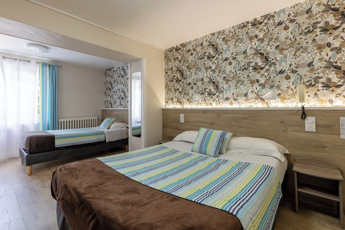 A hotel room with two beds, a wooden headboard, a floral-patterned wall, and a small bedside table.