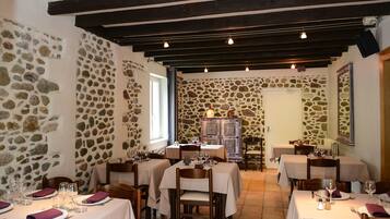 Un restaurante con paredes de piedra y vigas de madera, mesas dispuestas con manteles blancos y servilletas moradas, y una chimenea de piedra.