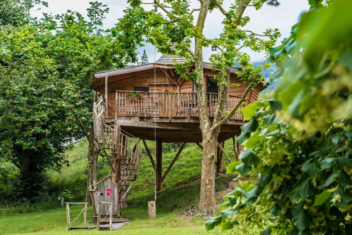 A wooden treehouse with a balcony, surrounded by lush greenery.