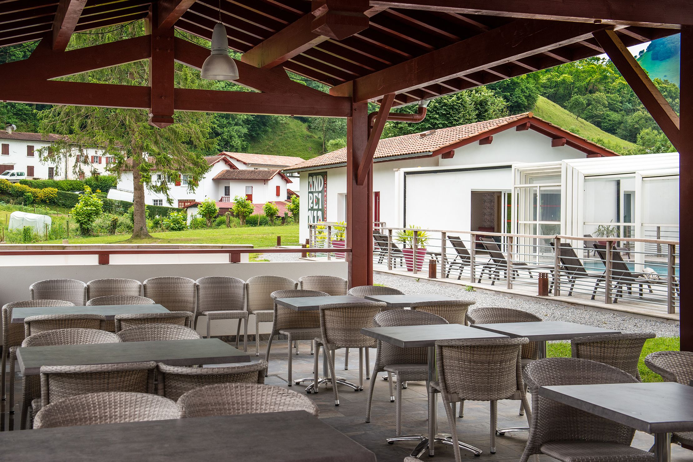 Un patio al aire libre con mesas y sillas de mimbre, una zona cubierta con vista a una piscina y casas al fondo.