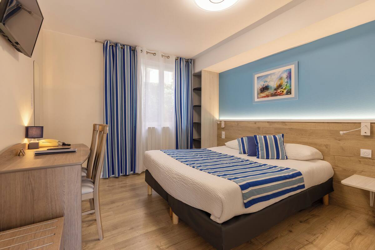 A modern hotel room with a wooden desk, a bed with striped bedding, a painting on the wall, and a view of the outdoors through the window.