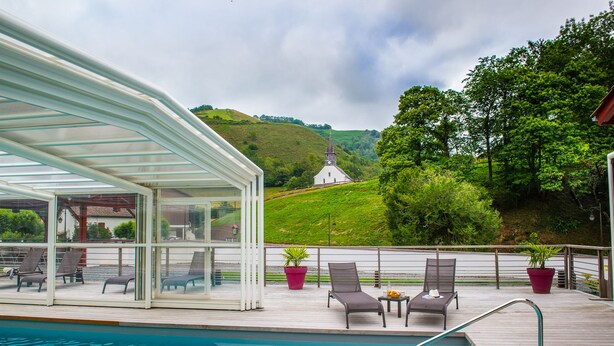A poolside area with a covered seating area, lounge chairs, and a view of a church on a hill. - Hôtel Andreinia (Esterencuby)