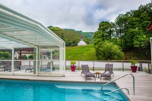 A poolside area with a covered seating area, lounge chairs, and a view of a church on a hill. - Hôtel Andreinia (Esterencuby)