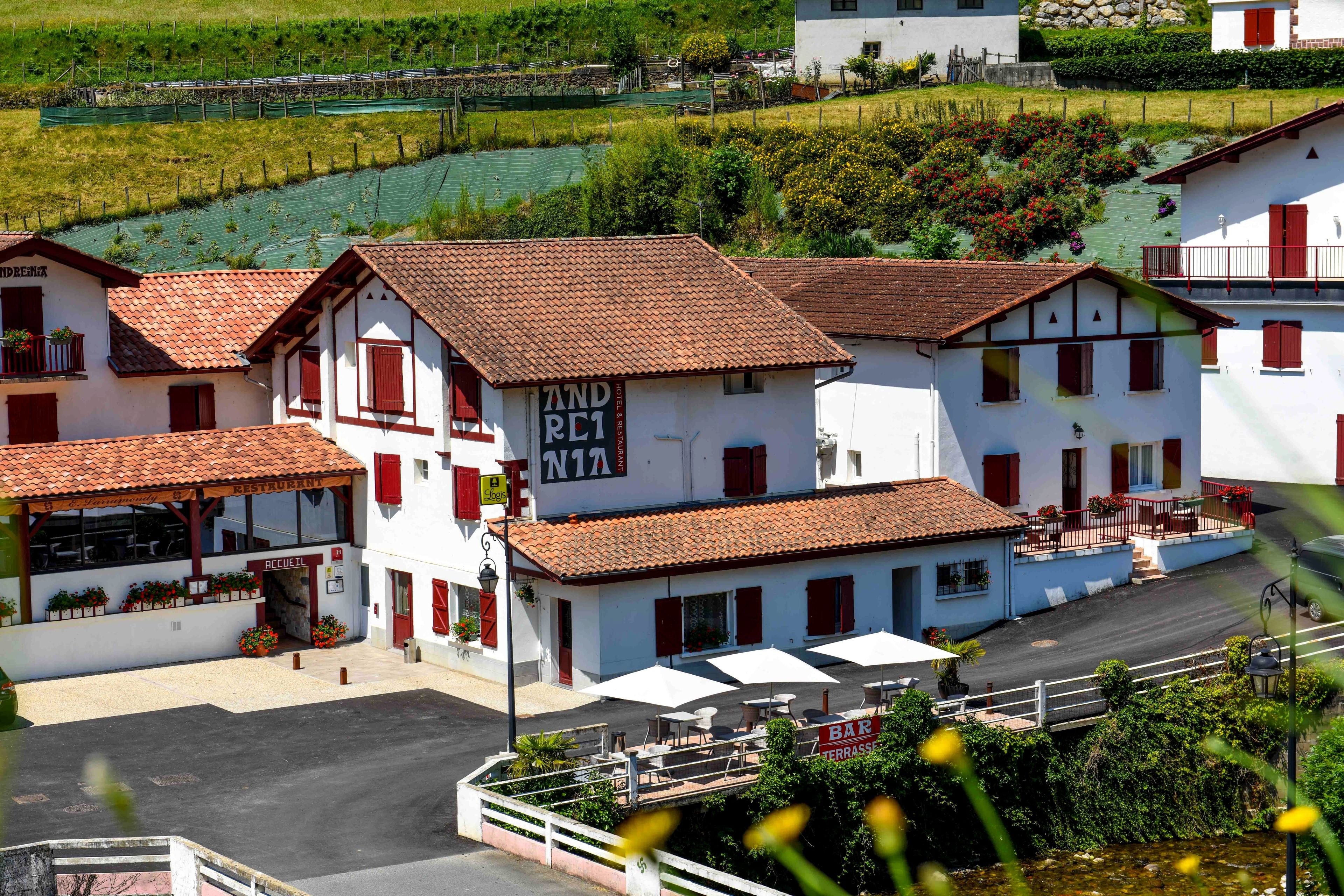 A white building with red shutters and a tiled roof, featuring a bar terrace with outdoor seating and umbrellas.