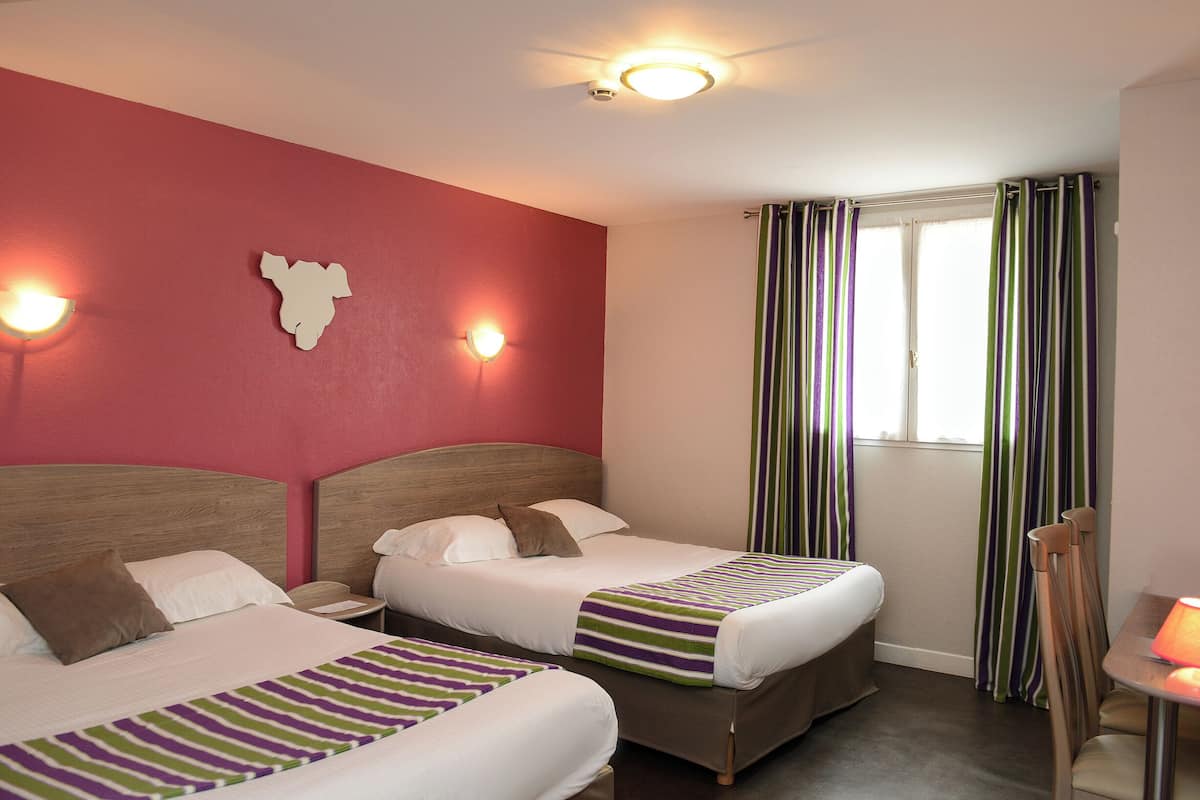 A hotel room with two beds, a red accent wall, a window with striped curtains, and a small table with a lamp.