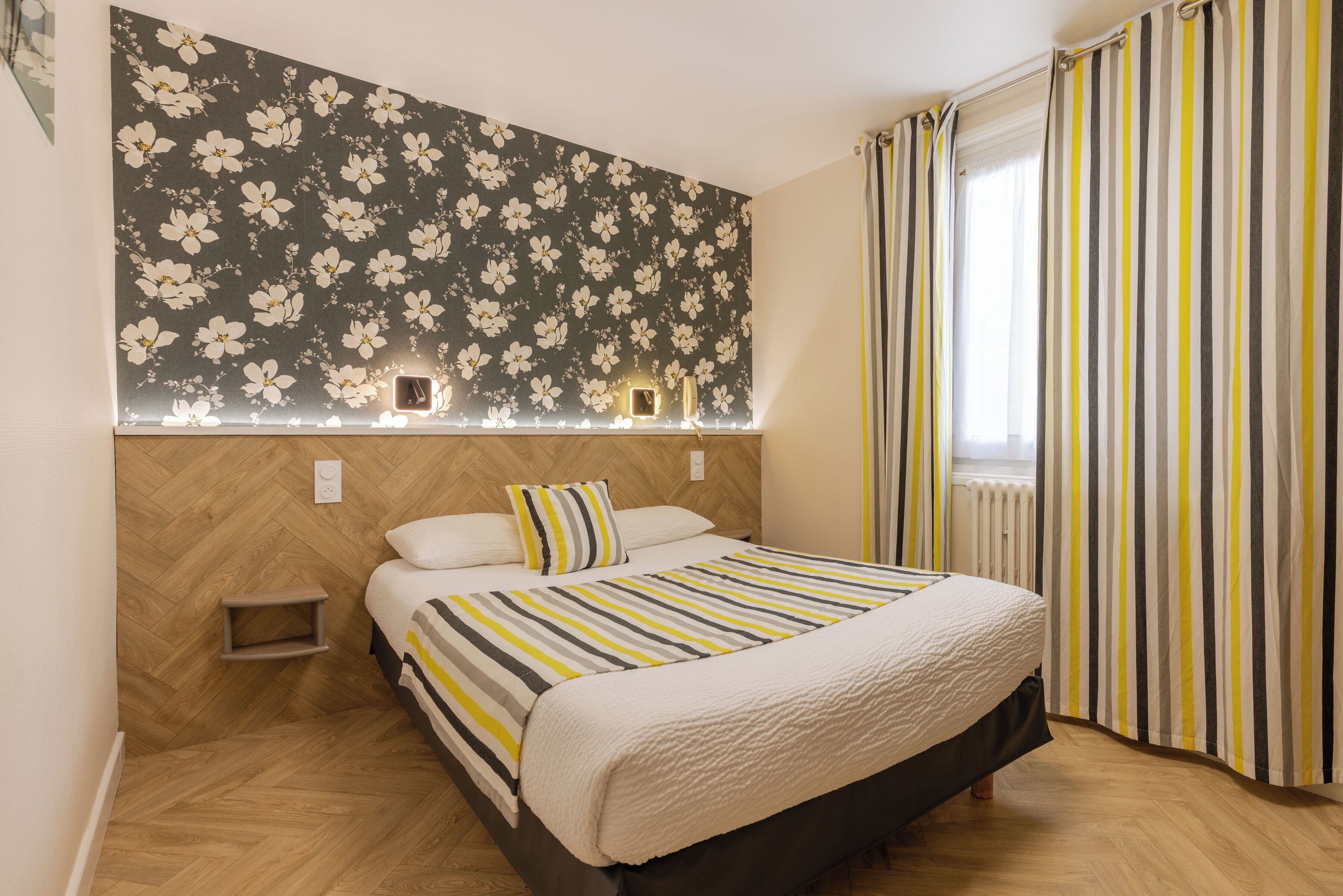 A bedroom with a bed, a nightstand, a wall-mounted light, and a window with curtains.
