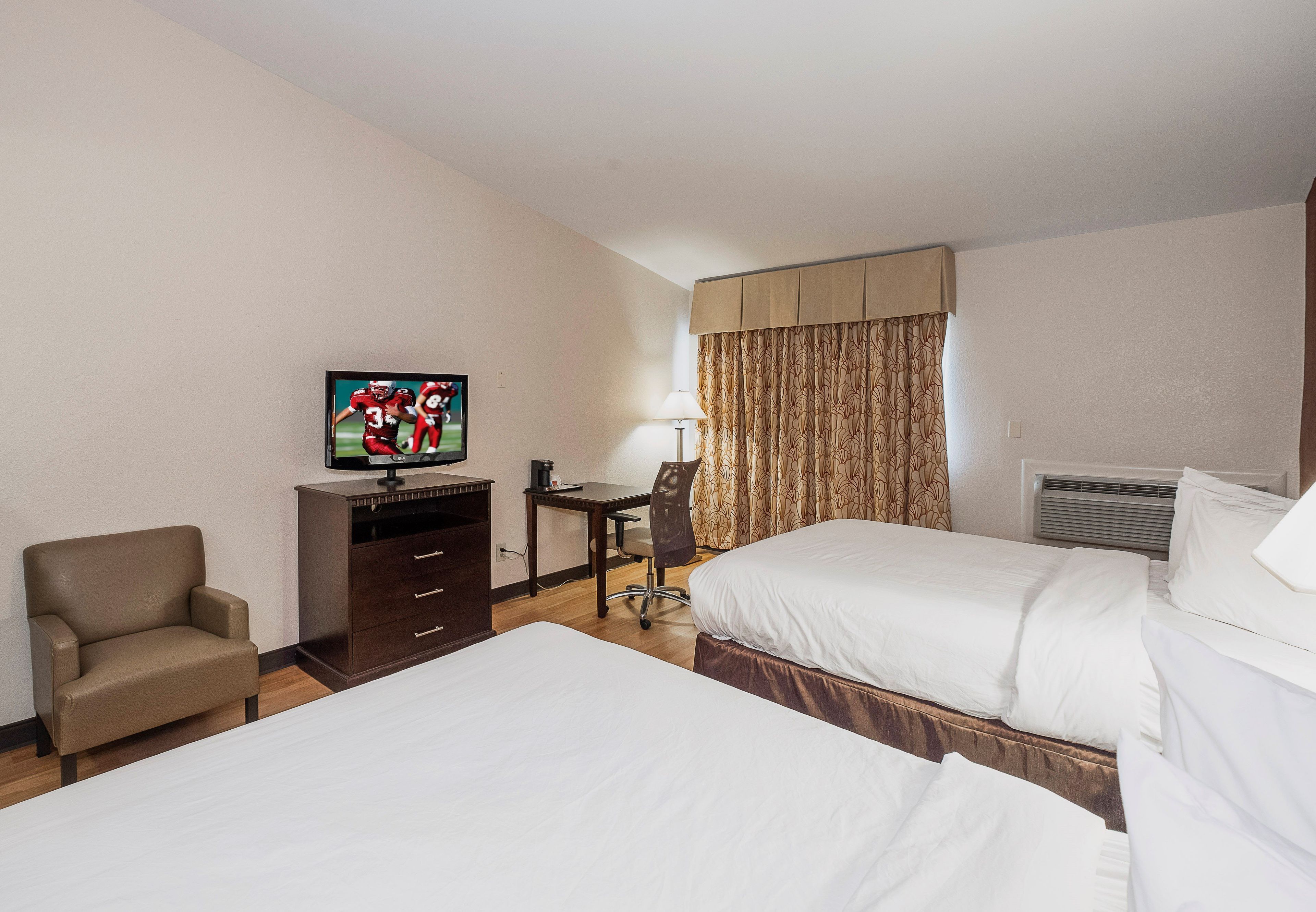 Quarto de hotel com duas camas, uma mesa, uma cadeira, uma televisão e uma janela com cortinas.
