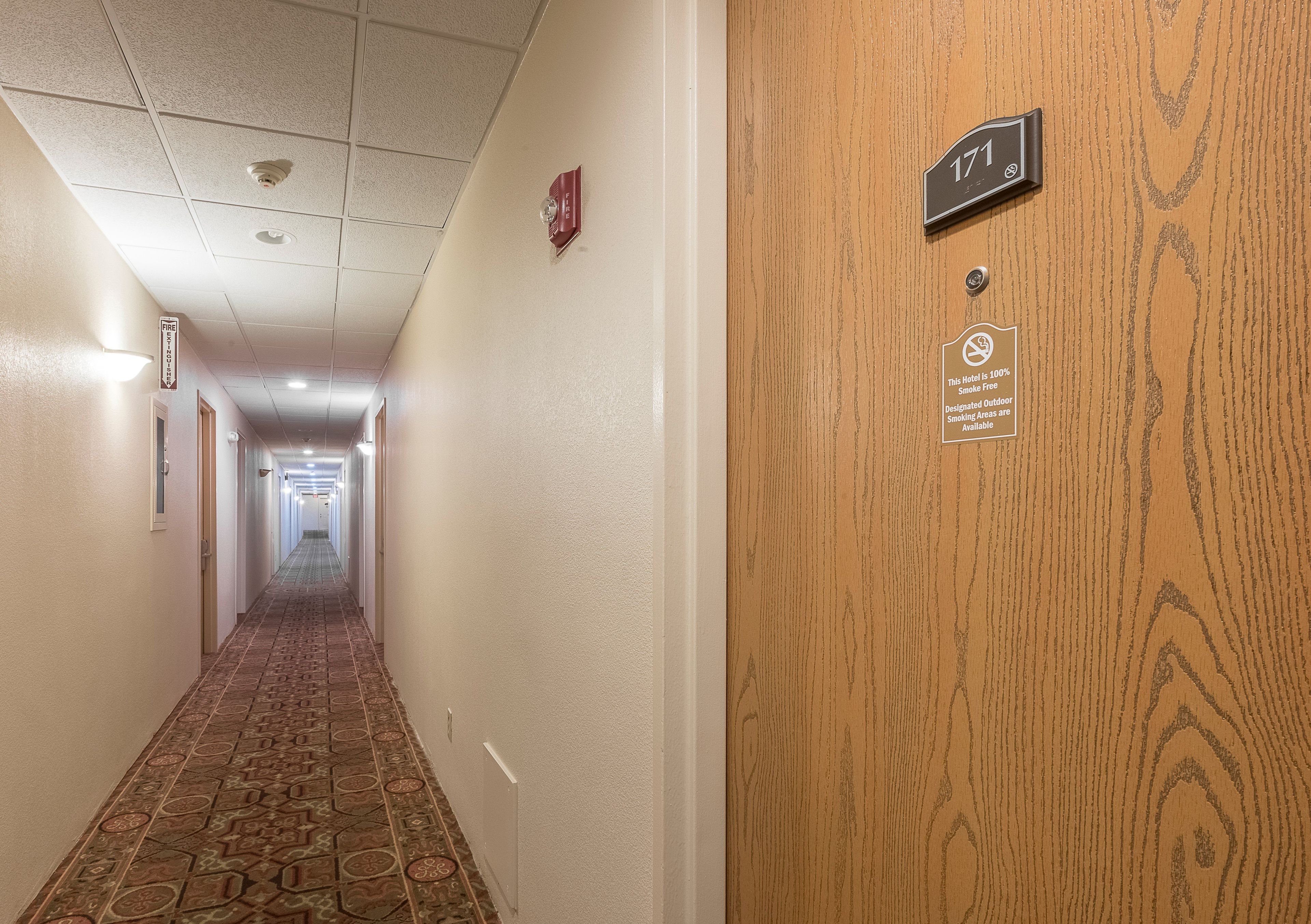 Corredor de hotel com sinal de saída de incêndio, detector de fumaça e placa indicando que é área livre de fumo na porta.