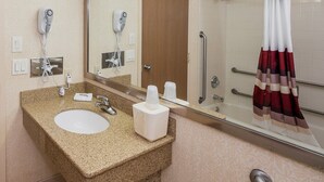 Un baño de hotel con encimera de granito, un lavamanos, un espejo, una ducha con puerta de cristal y un perchero con una toalla roja y blanca.