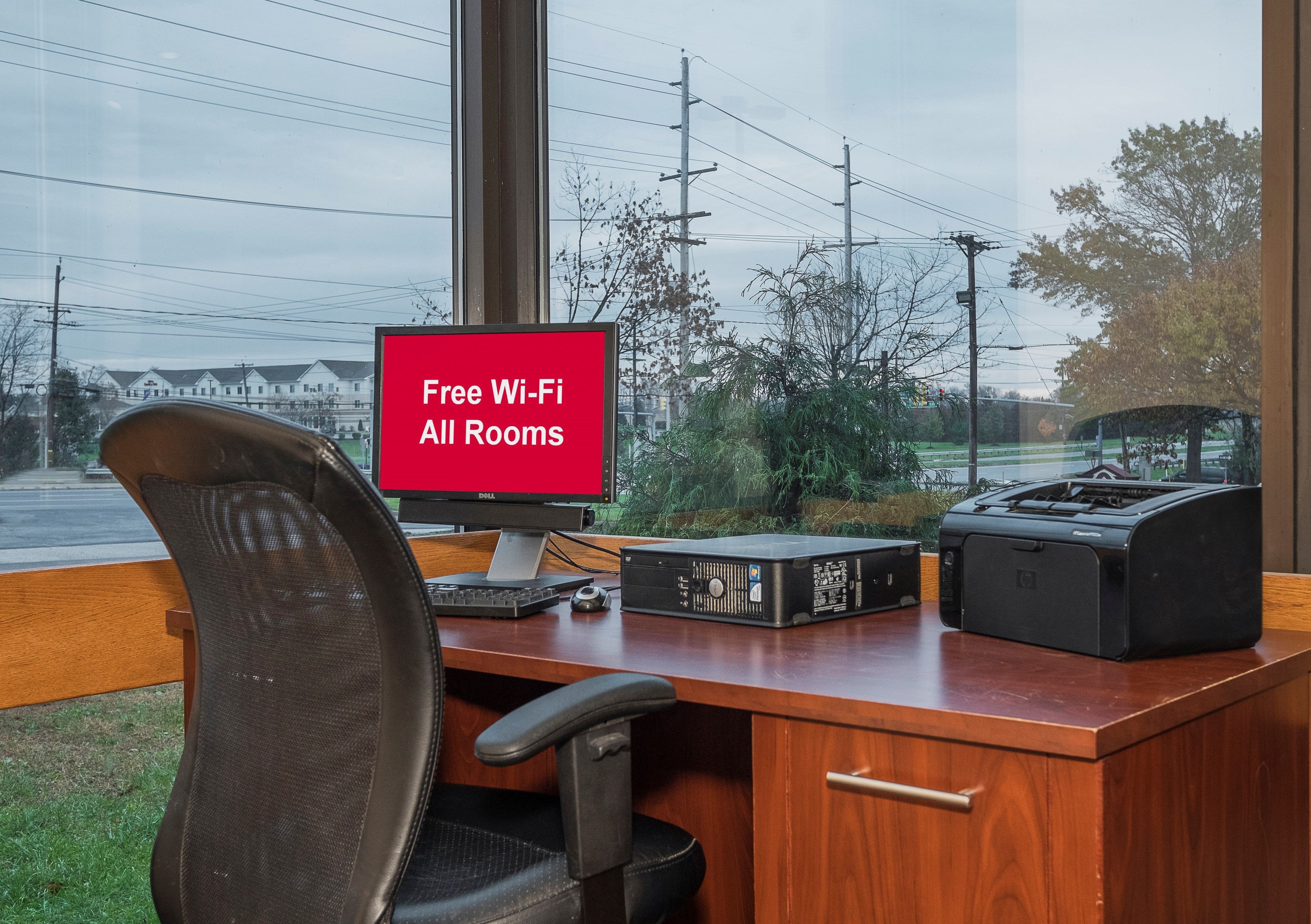 A hotel room with a desk, a computer, a printer, and a chair. A window with a view of the street and power lines is visible.