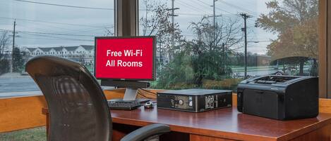 A hotel room with a desk, a computer, a printer, and a chair. A window with a view of the street and power lines is visible.