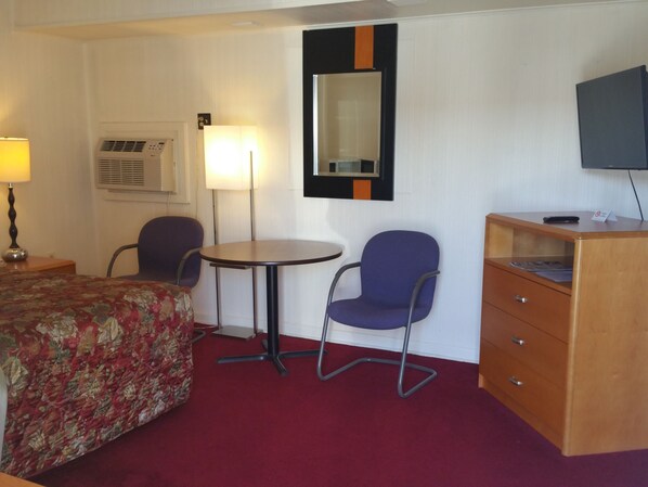 Una habitación de hotel con una cama, dos sillas, un escritorio, una lámpara, un espejo y un televisor.