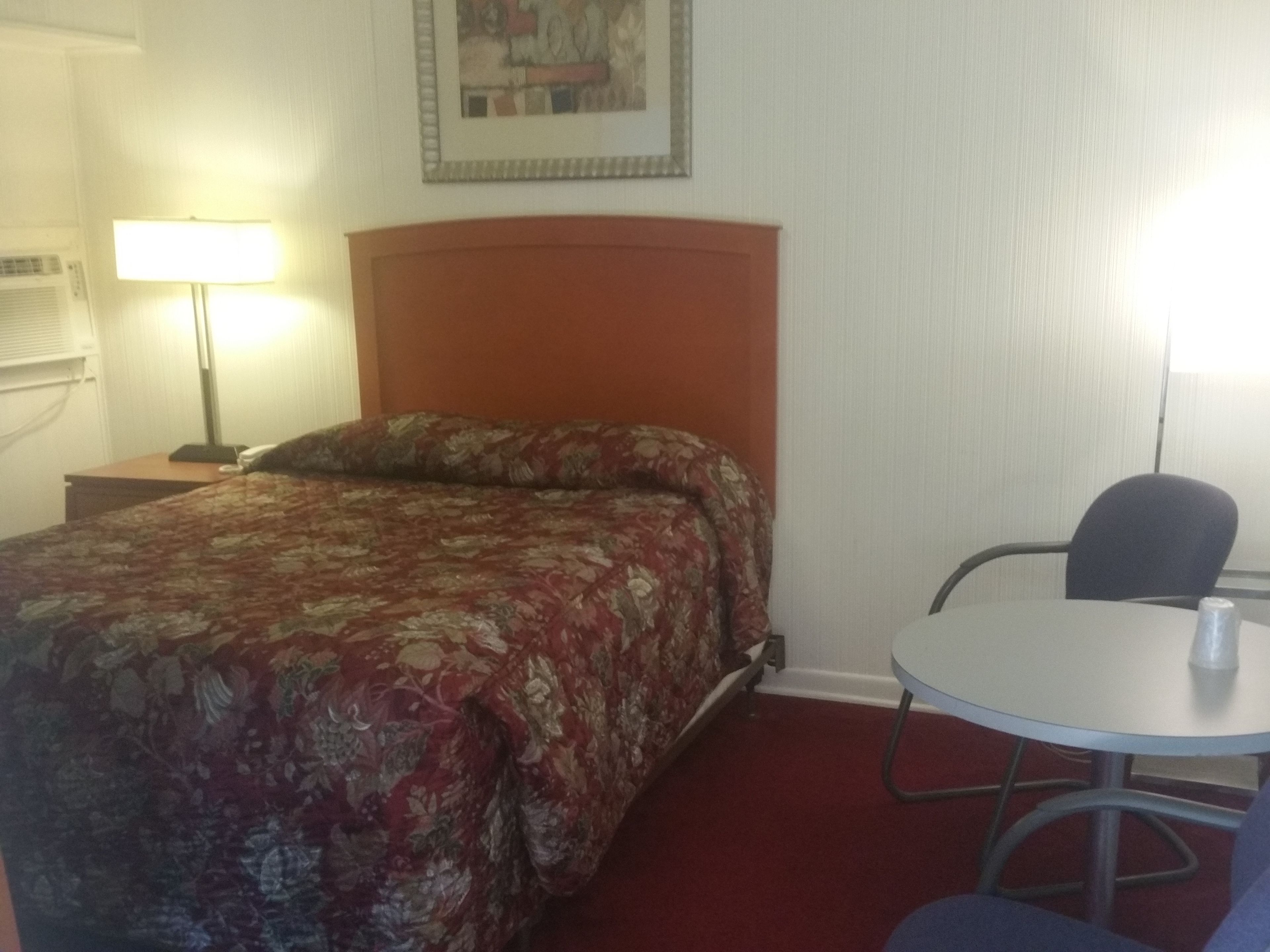 A hotel room with a bed, a bedside table with a lamp, a small round table with a chair, and a framed picture on the wall.