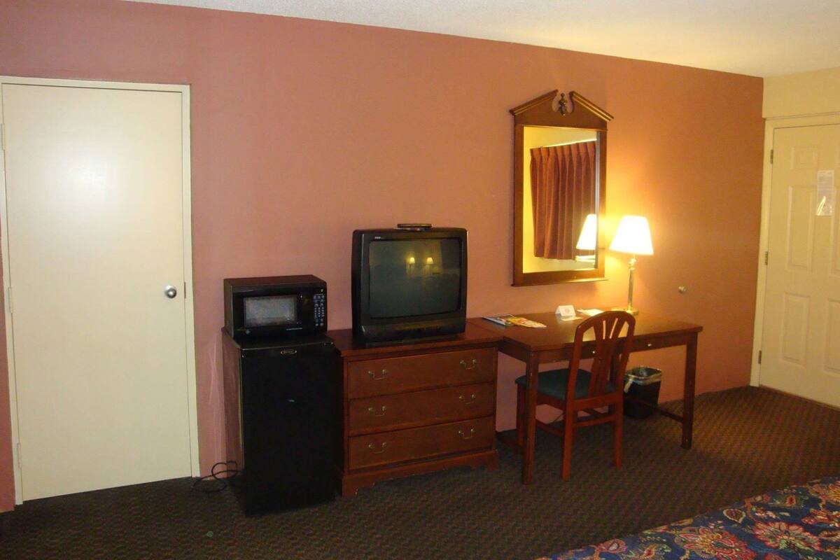 A hotel room with a television, a small desk with a chair, a dresser, a mirror, and a door.