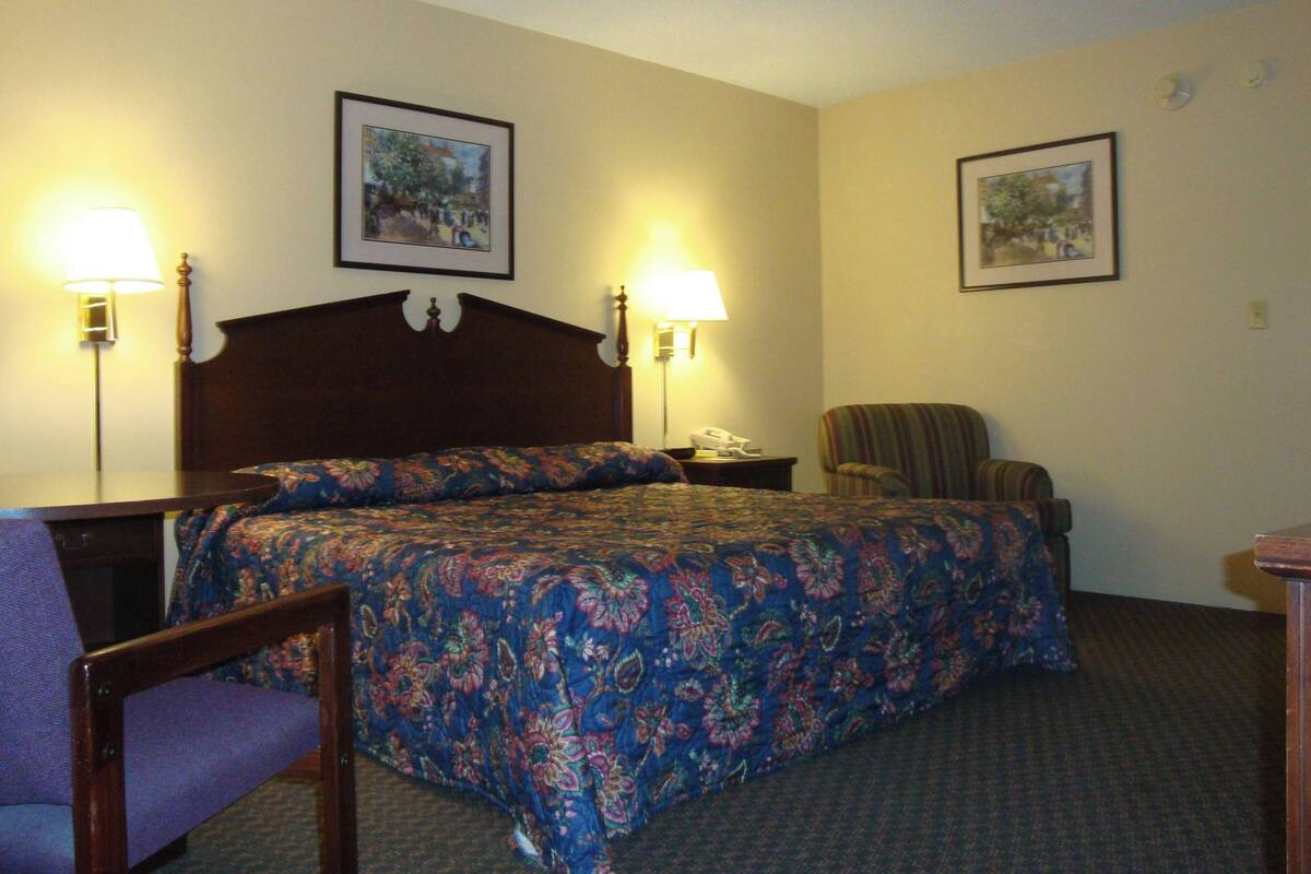 A hotel room with a bed, two bedside lamps, a chair, a desk, and two framed pictures on the wall.