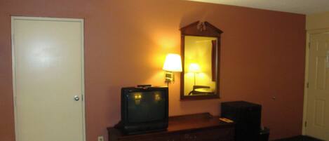 A hotel room with a closed door, a television on a wooden dresser, a mirror above the dresser, and a lamp on the wall.