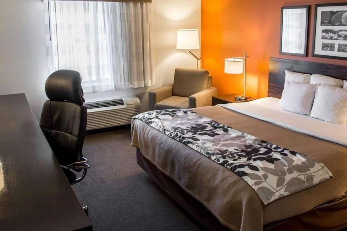 A hotel room with a bed, a desk, a chair, a window with curtains, a lamp, and framed pictures on the wall.