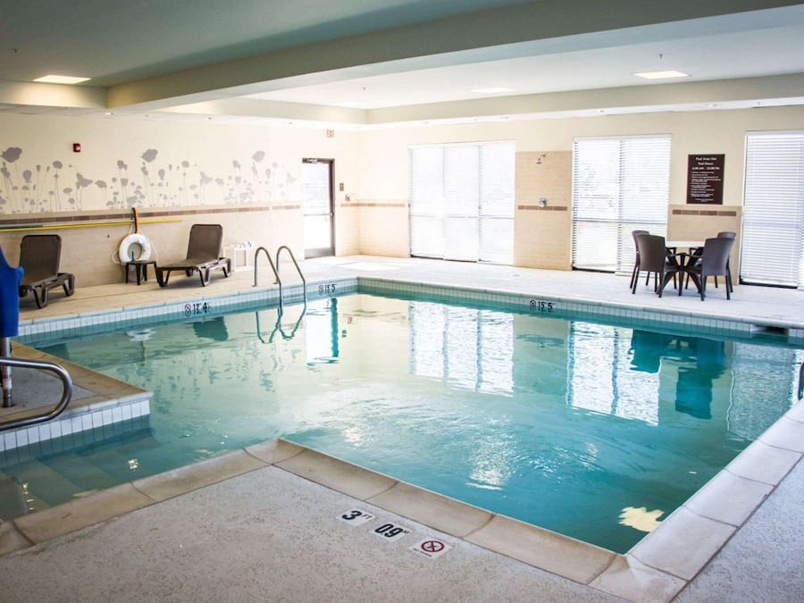 An indoor swimming pool with clear water, a lifeguard chair, lounge chairs, and a poolside seating area with tables and chairs.