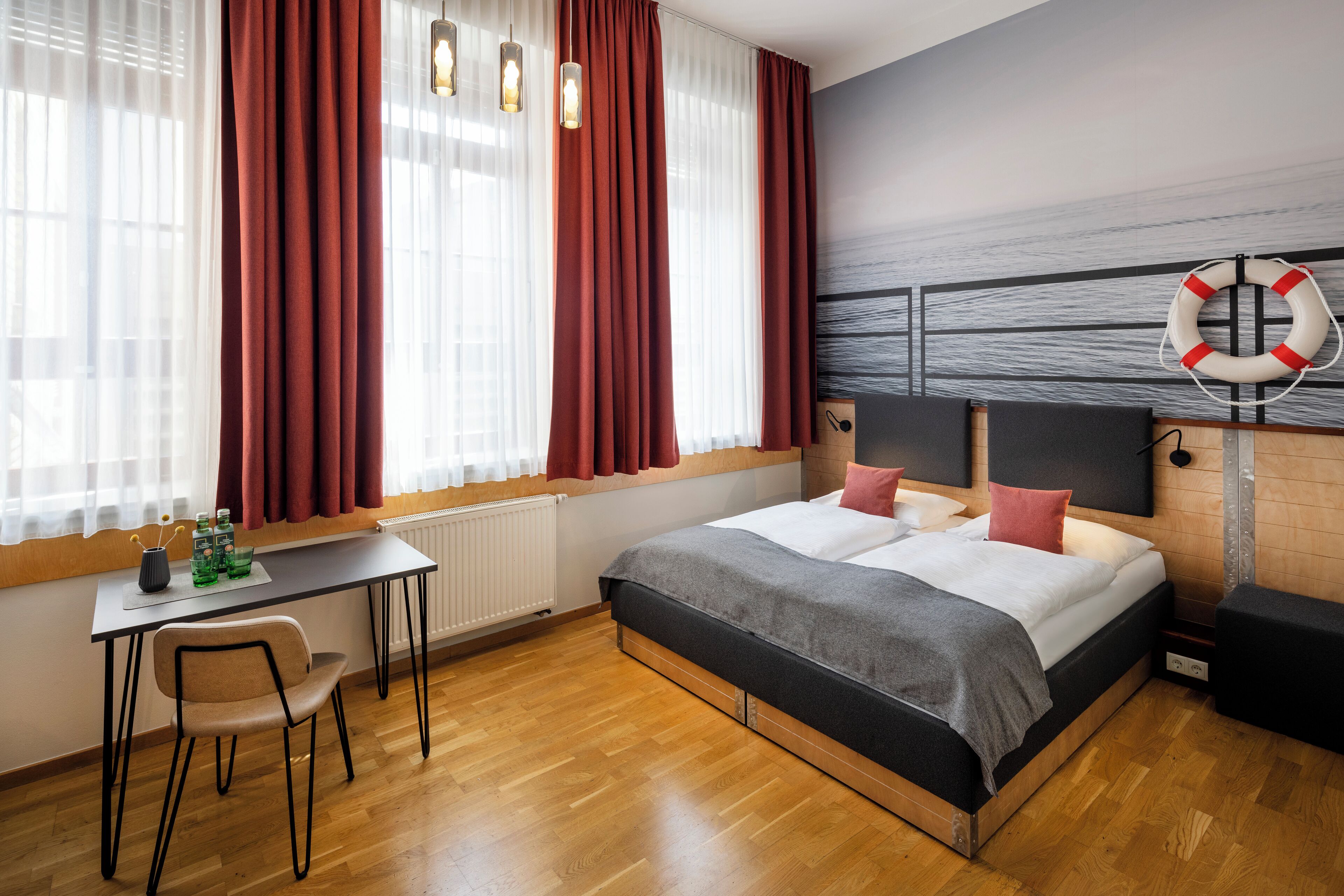 A modern hotel room with a large bed, a desk, and a chair. There is a window with curtains and a wall-mounted life preserver.
