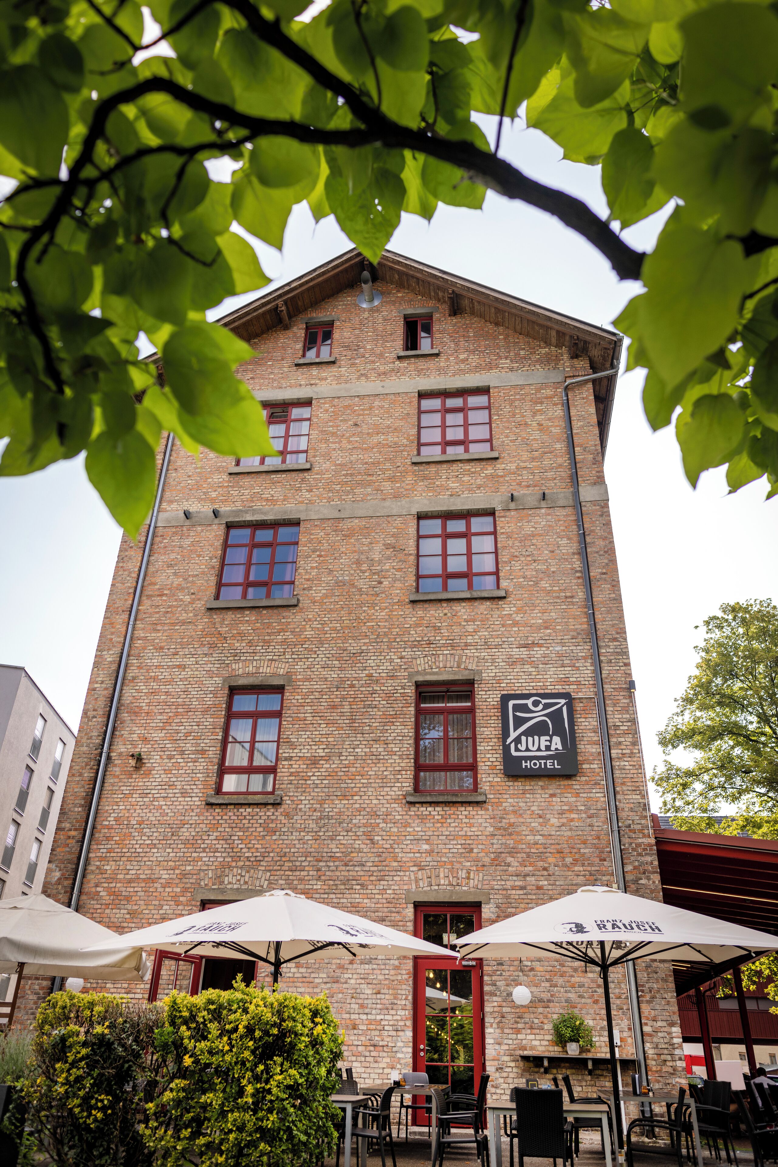 Ein Gebäude aus Backsteinen mit einem Schild für das JUFA Hotel. Unter Sonnenschirmen stehen Tische und Stühle im Freien.