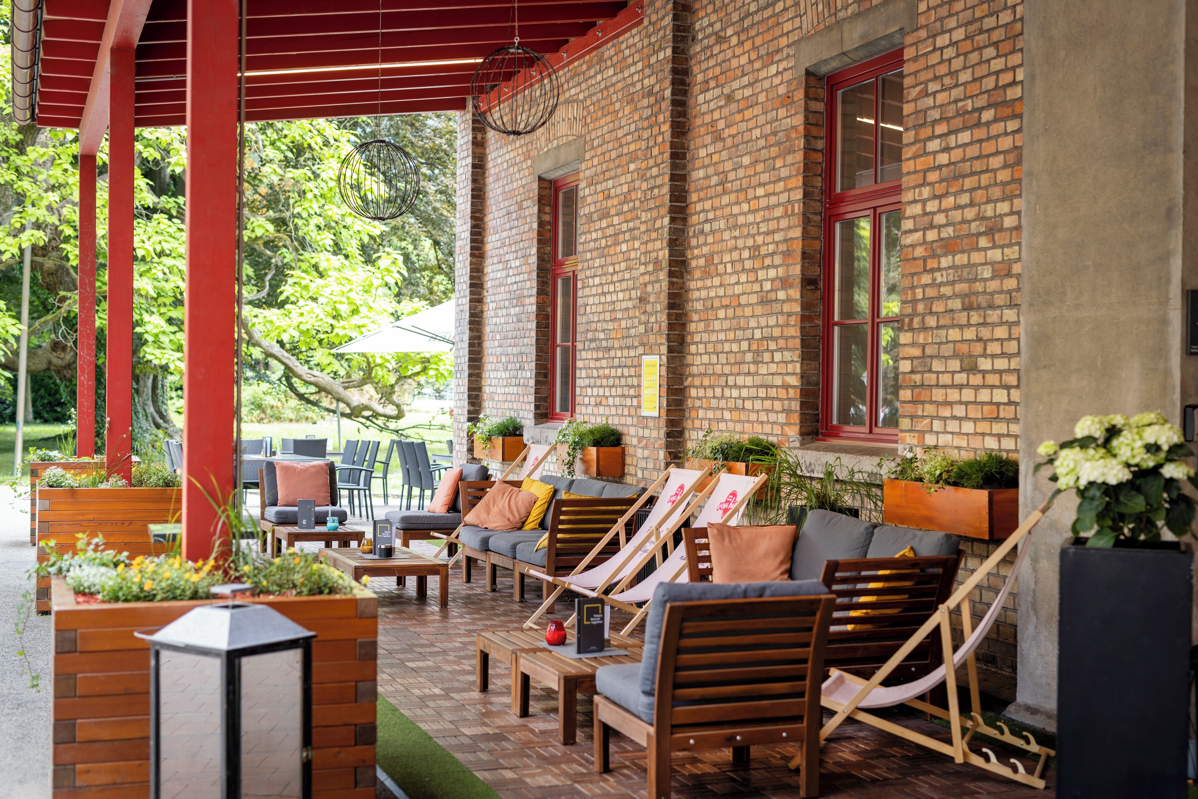 Un espace extérieur aménagé avec des chaises longues en bois et des jardinières, un bâtiment en briques et une partie couverte avec des poutres rouges.