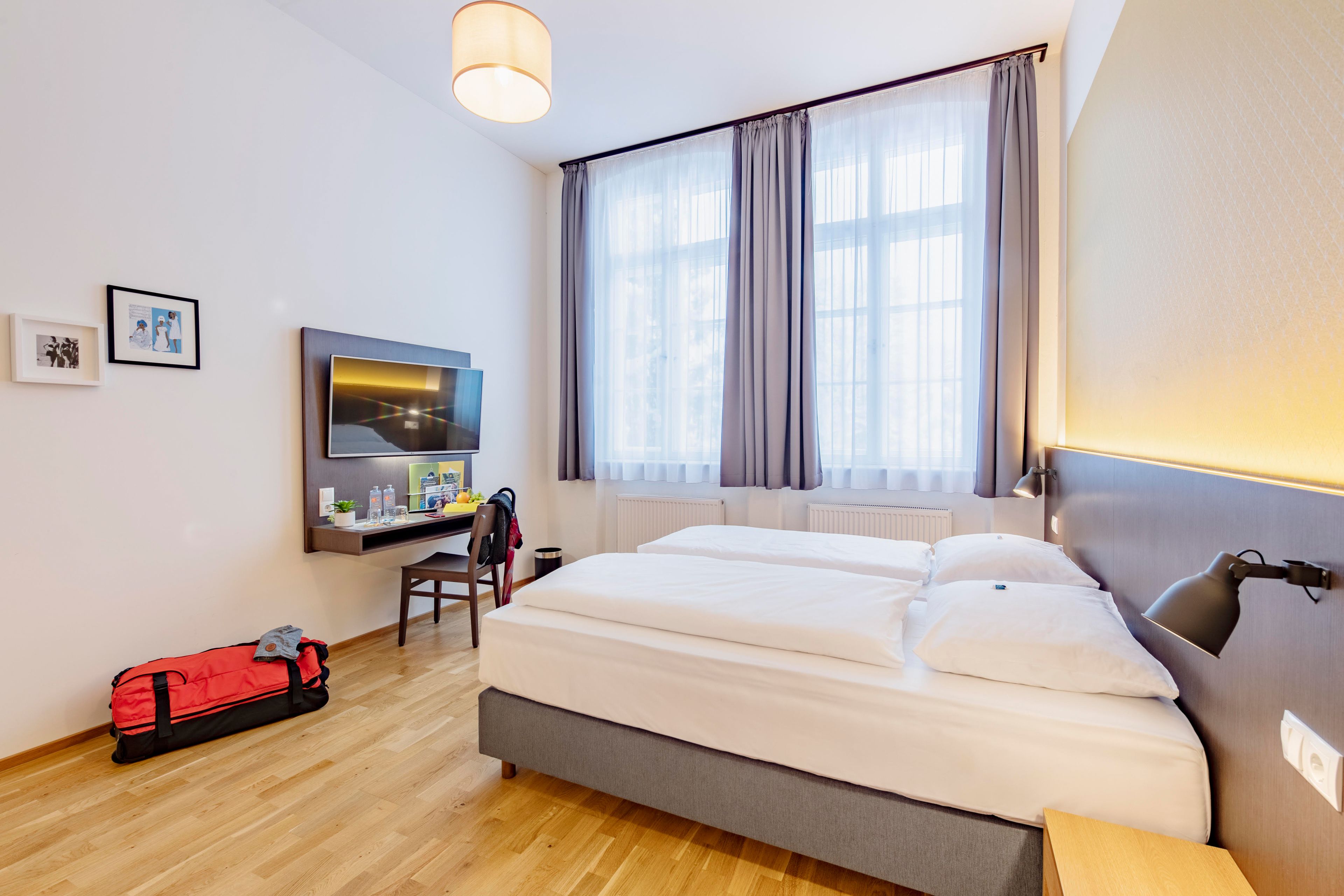 A hotel room with a bed, a desk with a computer, a chair, a red suitcase, and a window with curtains.