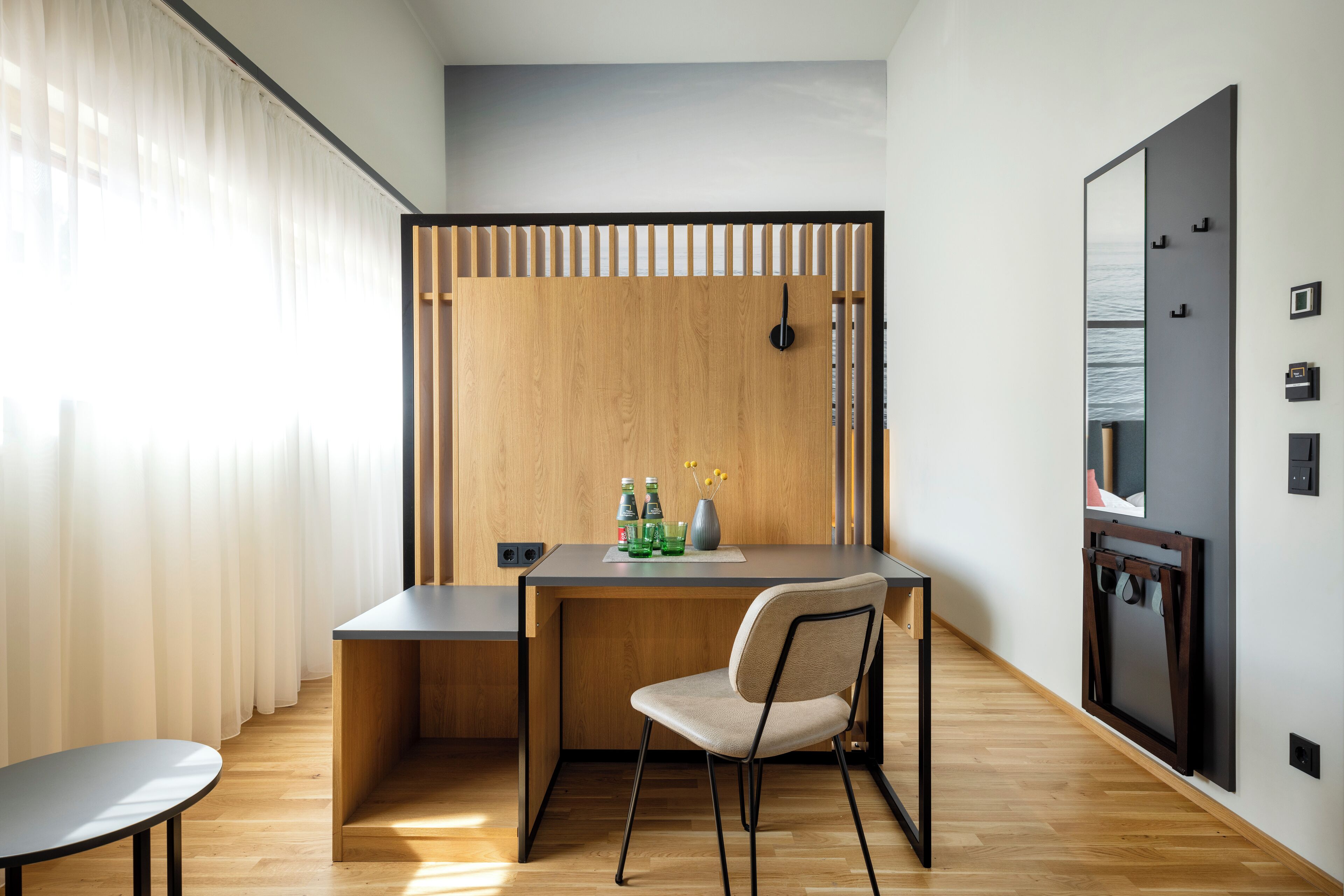 Une pièce moderne avec un bureau en bois, une chaise et une petite table. On y trouve une fenêtre avec des rideaux légers, une étagère fixée au mur ornée d’objets décoratifs et un tableau encadré.