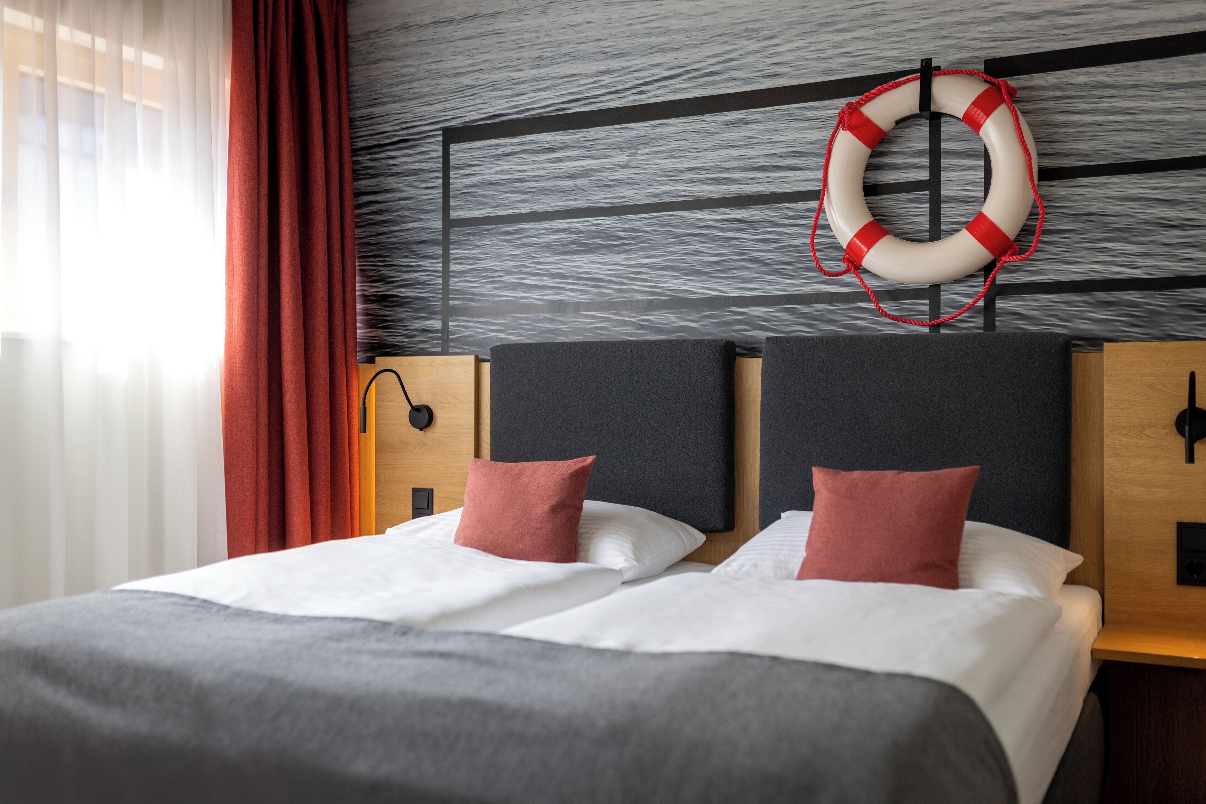 Ein Doppelbett mit weißen Leinen und zwei roten Kissen, ein Rettungsring an der Wand und ein Holz-Kopfteil.