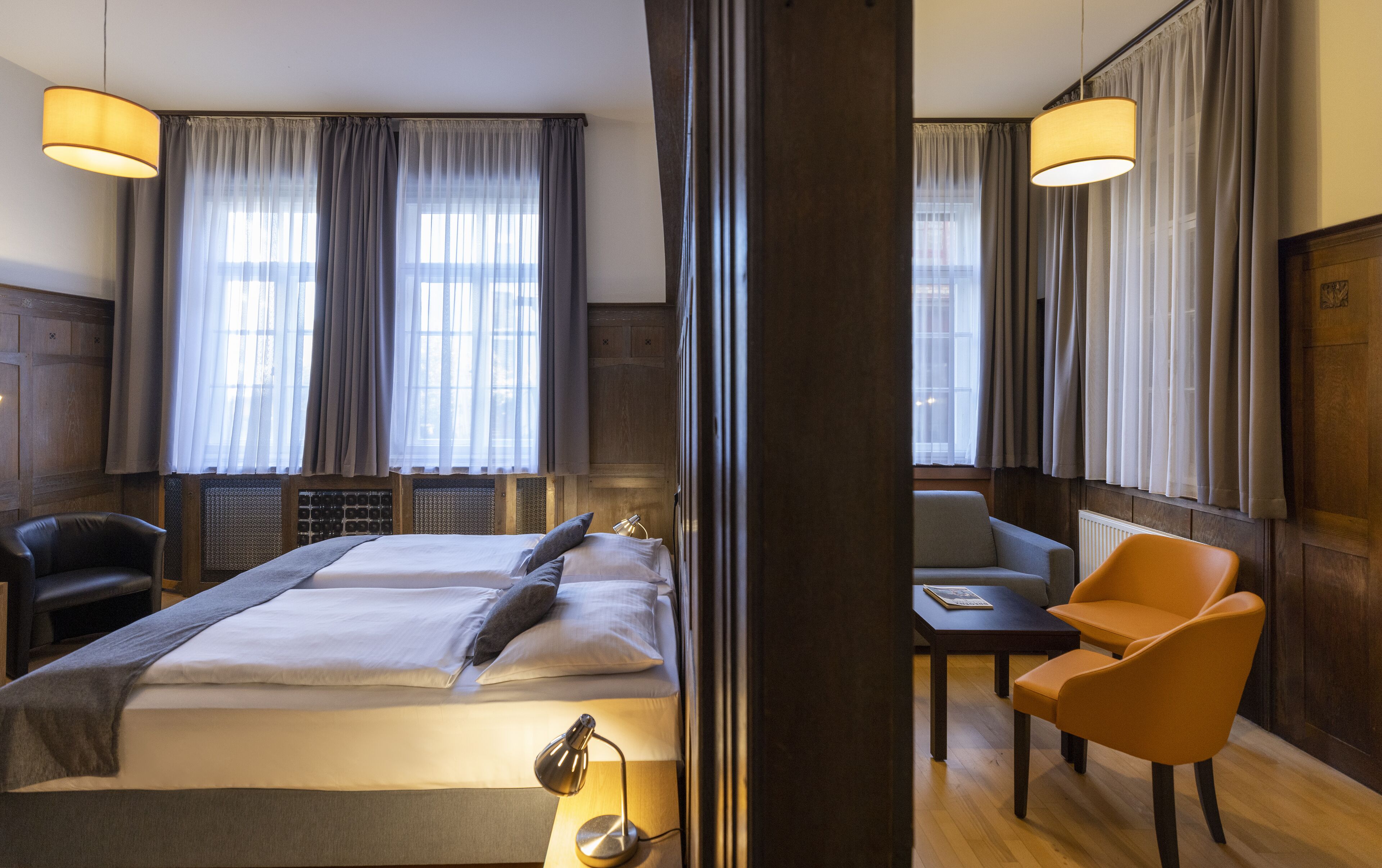 Une chambre d’hôtel avec un lit, deux chaises, une table et de grandes fenêtres avec des rideaux.