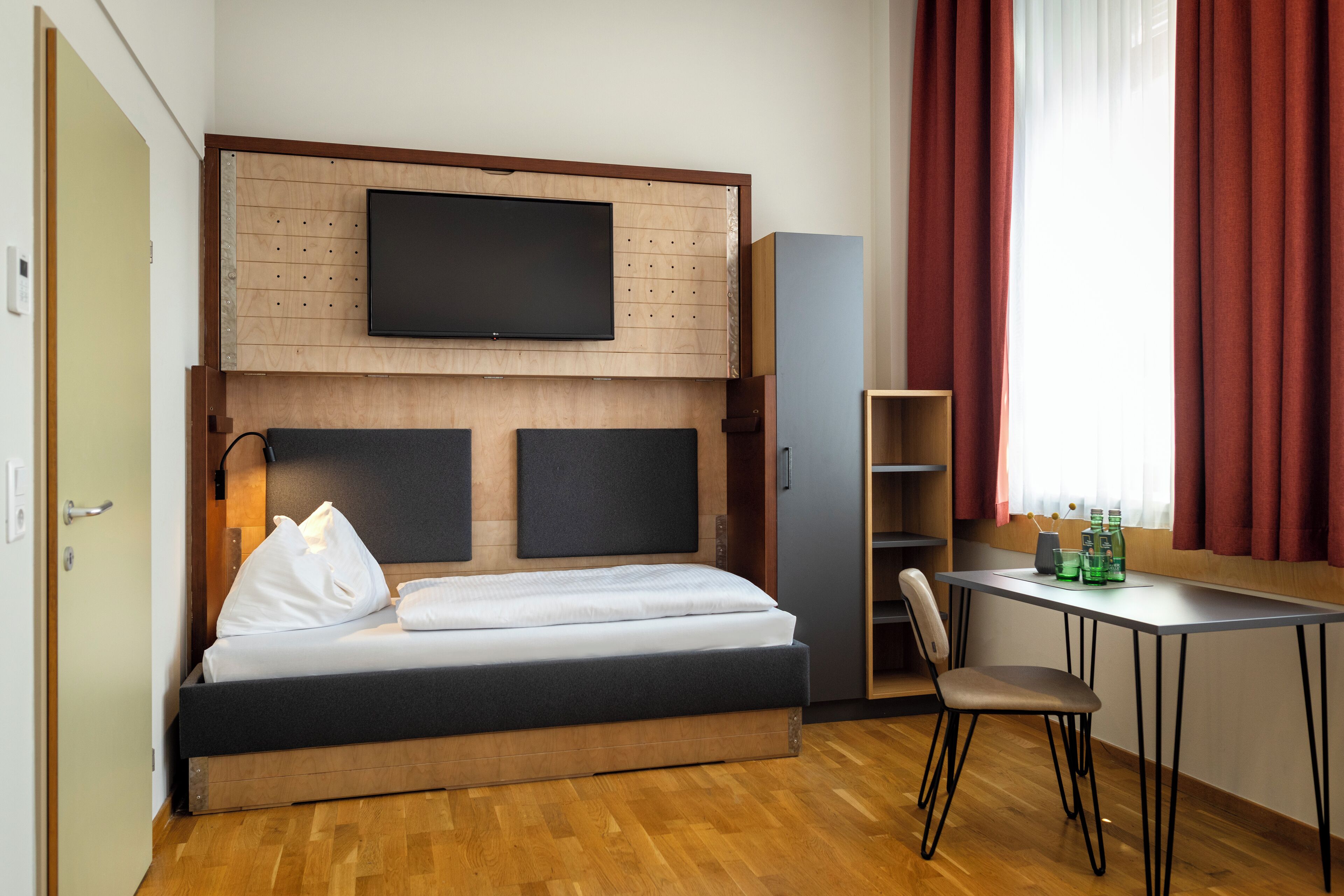 A hotel room with a bed, a TV mounted on the wall, a desk with a chair, and a window with curtains.