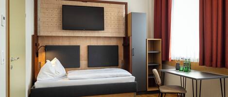 Una habitación de hotel con cama, televisión montada en la pared, escritorio con silla y ventana con cortinas.