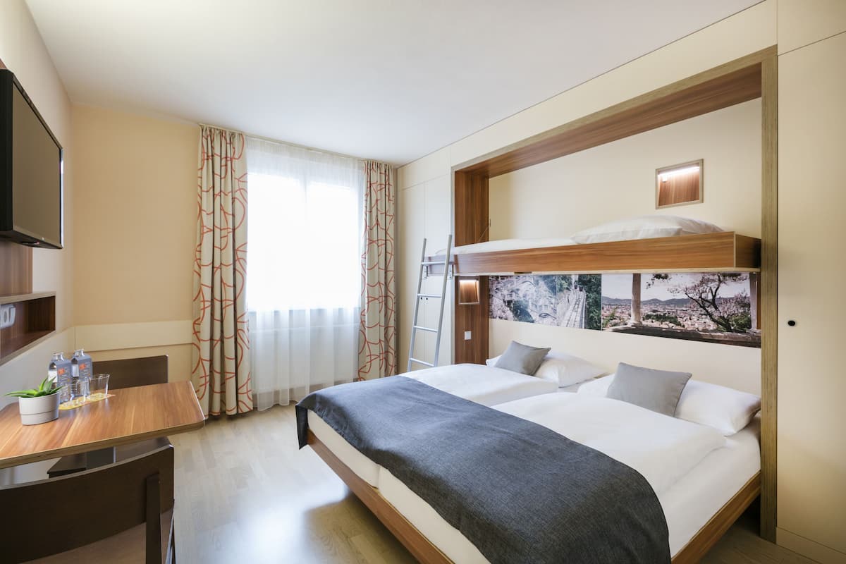 A hotel room with a bunk bed, a desk with a chair, a TV, and a window with curtains.