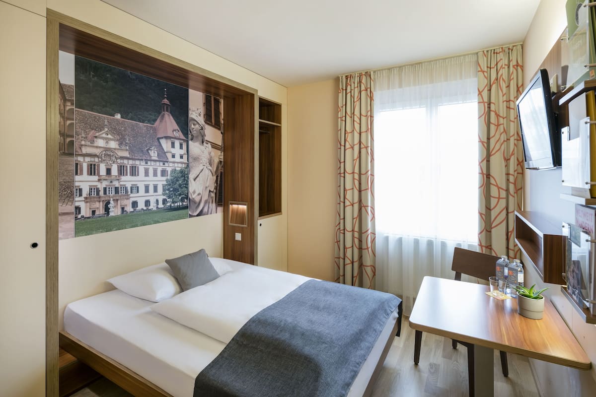 A hotel room with a bed, a table, a TV, and a painting of a building.