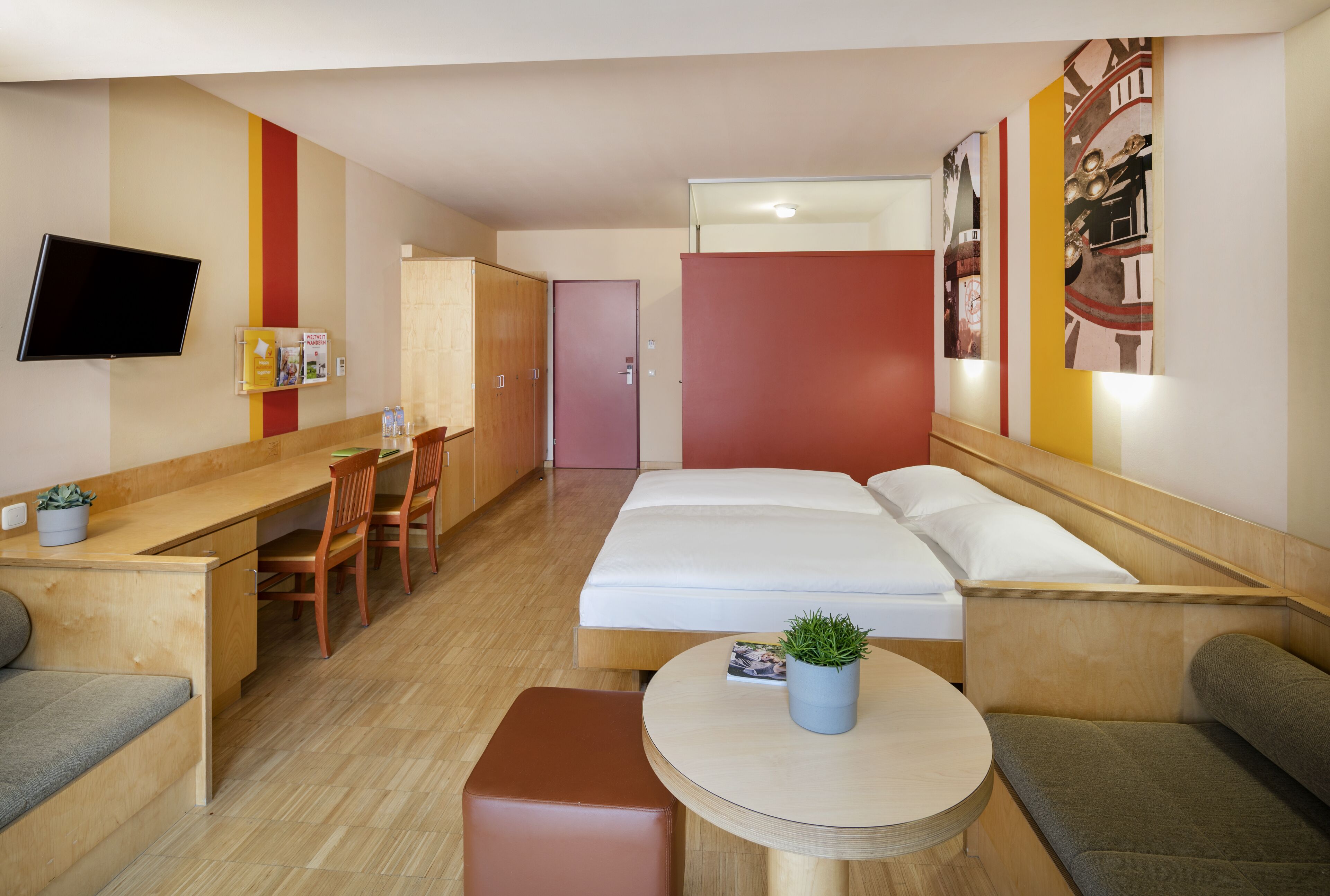 A hotel room with a bed, a desk, a chair, a small table, and a TV.