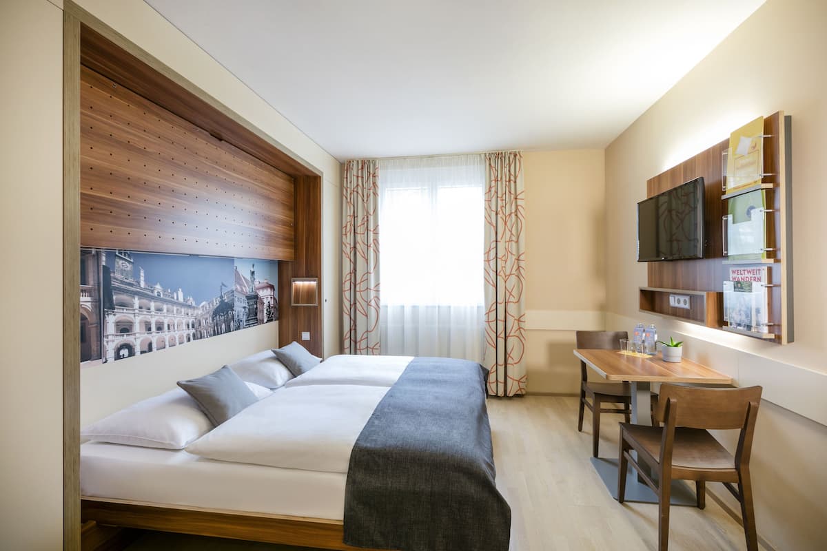 A hotel room with a bed, a dining table with chairs, a TV mounted on the wall, and a window with curtains.